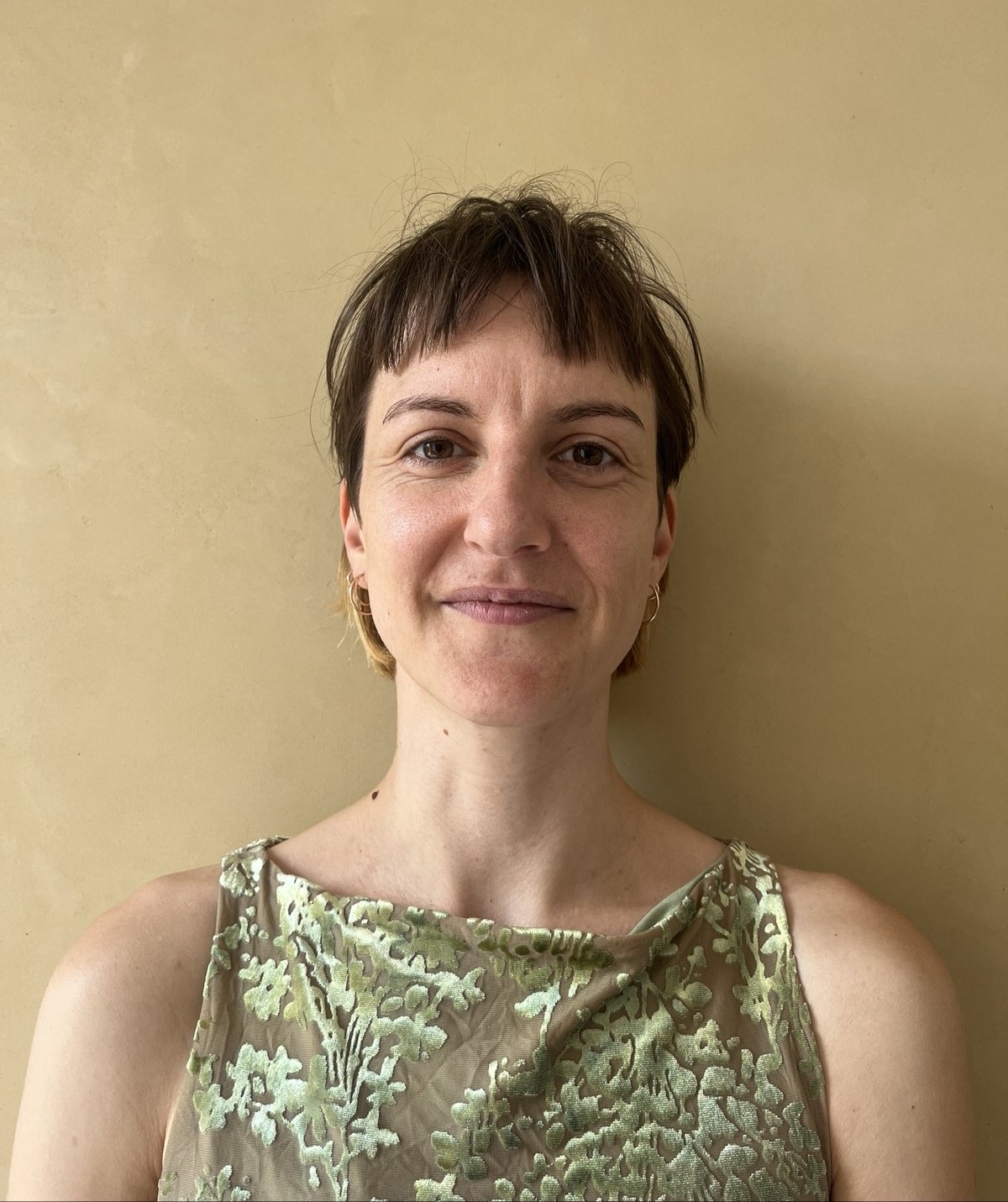 Woman with short brown hair in a green floral sleeveless top, smiling against a beige wall.Head-and-shoulders portrait with a gentle smile and hoop earrings.