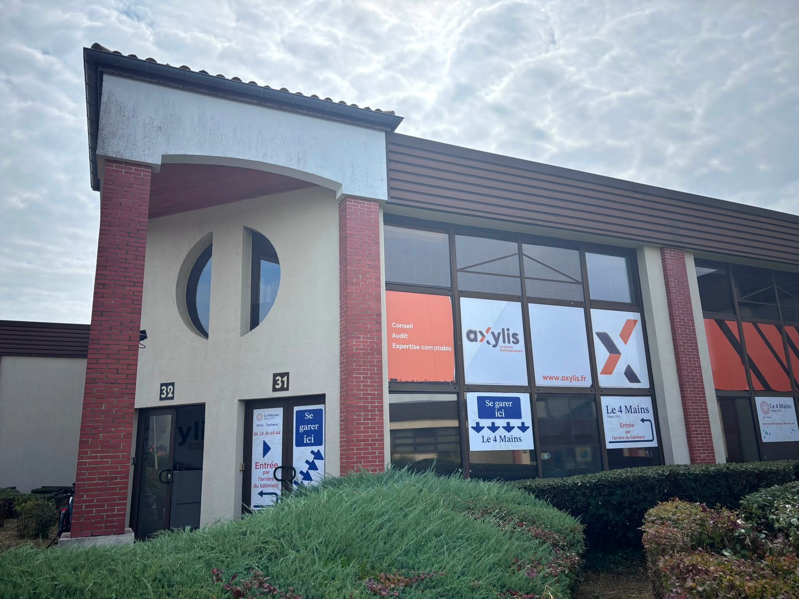 Exterior of a modern brick-and-stucco office building with large glass storefront windows and banners with orange and blue signage (axylis.fr).